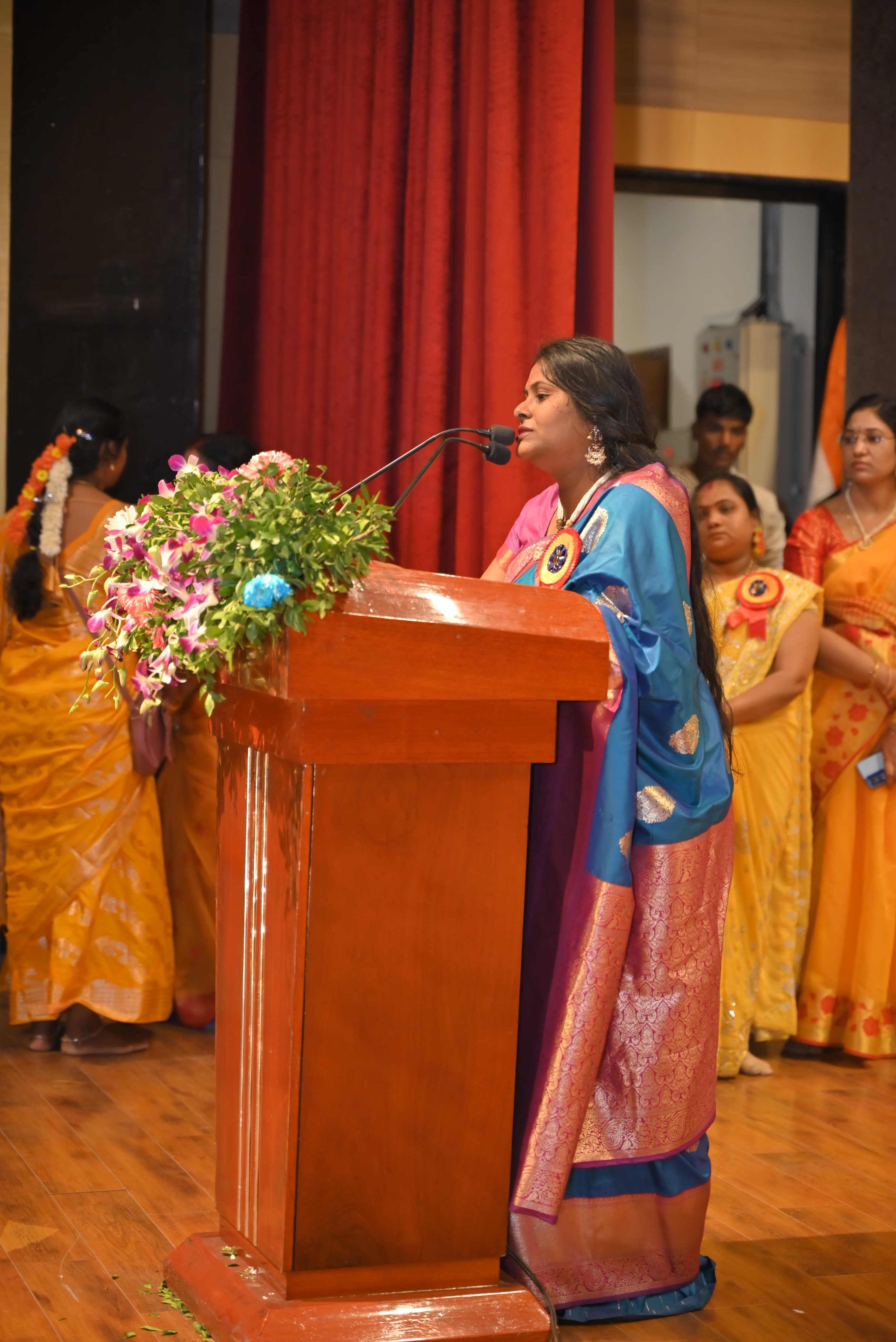 A woman in a blue sari speaking from a flower-decorated podium